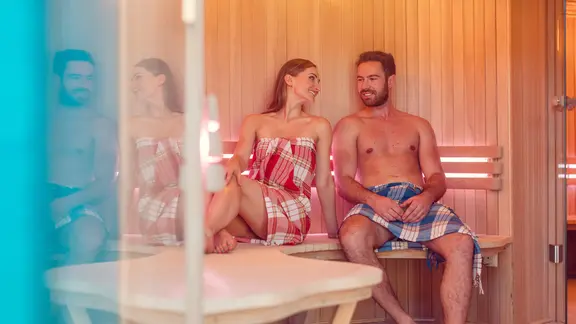 Junge Frau und junger Mann sitzen mit einem Tuch umwickelt in der Sauna.