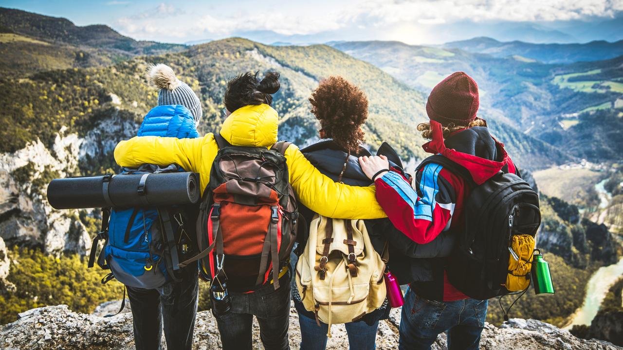Vier junge Menschen mit Rucksack stehen Arm in Arm an einem Felsen vor Bergpanorama.