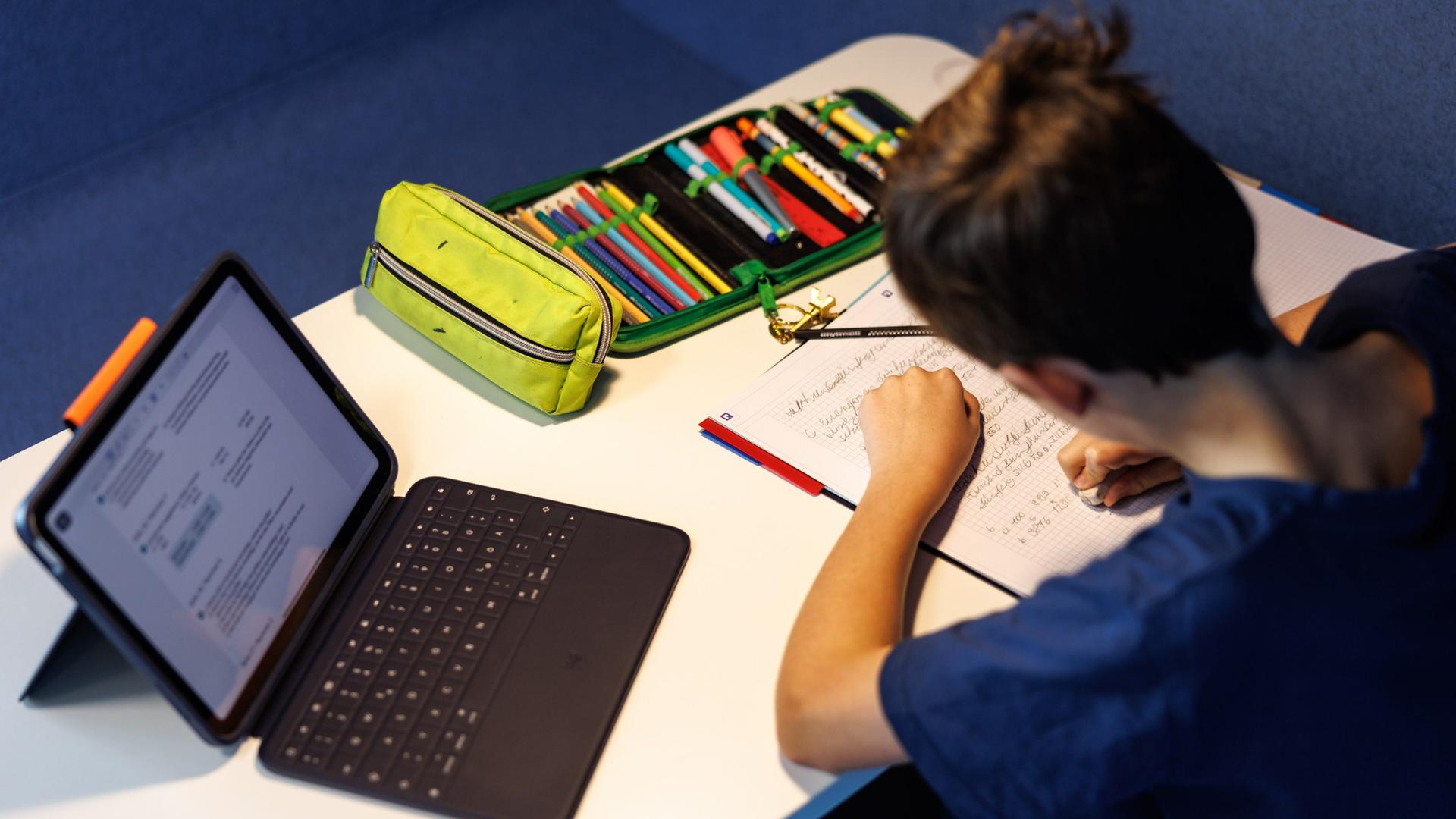 Ein Schüler einer fünften Klasse macht sich Notizen, während neben ihm ein Tablet zu sehen ist. | dpa, Matthias Balk