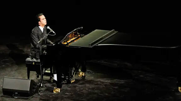 Nick Cave sitzt im schwarzen Anzug an einem Konzertflügel auf der Bühne der Elbphilharmonie. Er singt ins Mikrofon, während er Klavier spielt. Die Bühne ist dunkel ausgeleuchtet, der Fokus liegt ganz auf ihm und dem Flügel.