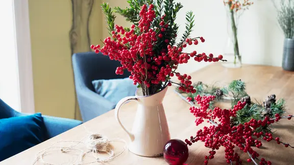 Eine Vase mit roten Ilex-Beeren und grünen Myrte-Zweigen steht auf einem Tisch.