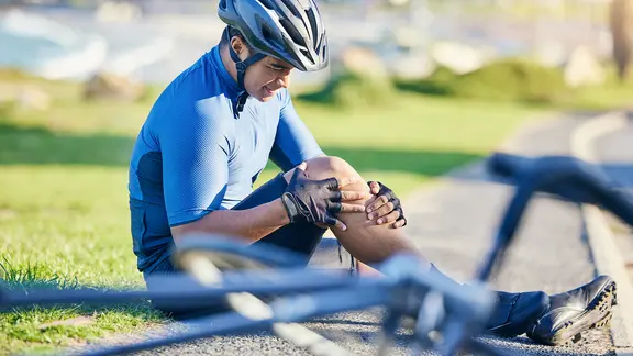 Ein Mann mit Helm sitzt auf dem Boden und hält sich sein Bein, vor ihm ein umgestürztes Fahrrad.