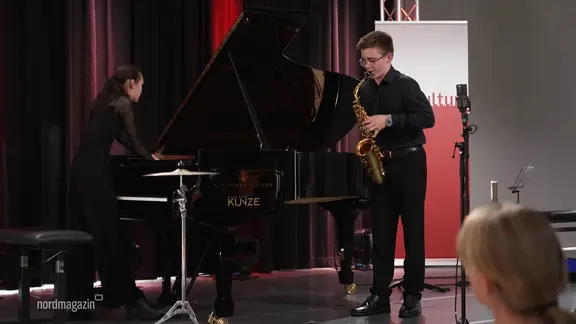 Ein Jugendlicher spielt auf einer Bühne Saxophon während er von einer Pianistin begleitet wird.