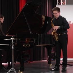 Ein Jugendlicher spielt auf einer Bühne Saxophon während er von einer Pianistin begleitet wird.