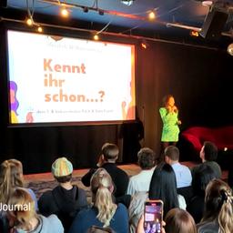 Vor einer Bühne mit Leinwand sitzen zahlreiche Personen und lauschen einer Präsentation.