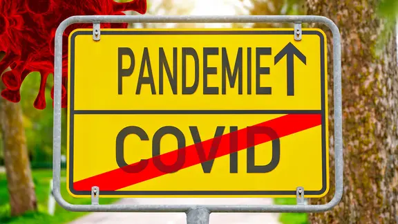 Auf einem Ortsschild ist das Wort "Covid" durchgestrichen, ein Pfeil weist Richtung Pandemie. (Fotomontage)