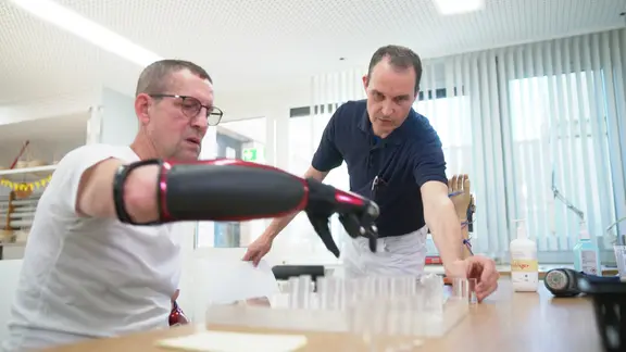 Ein Mann mit Hand-Prothesen übt, kleine Plastikröhrchen zu bewegen, die vor ihm auf einem Tisch stehen. 