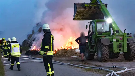 Brand in Hagermarsch: Mehrere Feuerwehrleute stehen vor dem Feuer. Ein Trecker hilft der Feuerwehr.