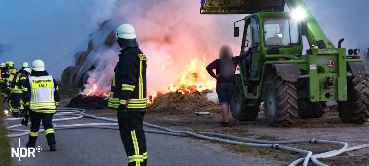 Der Gebäude-Rohbau und das Strohlager liegen laut Polizei nah beieinander. Die Beamten gehen von Brandstiftung aus. | Nonstopnews Brand in Hagermarsch: Mehrere Feuerwehrleute stehen vor dem Feuer. Ein Trecker hilft der Feuerwehr.