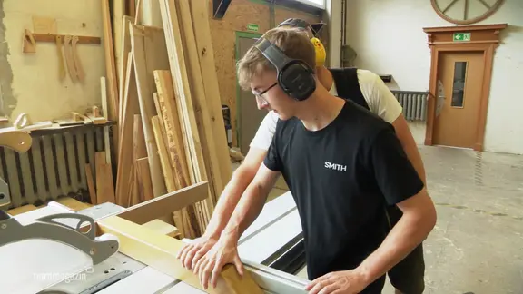 Ein Tischler hilft einem Praktikanten beim Zuschneiden eines Holzstücks.