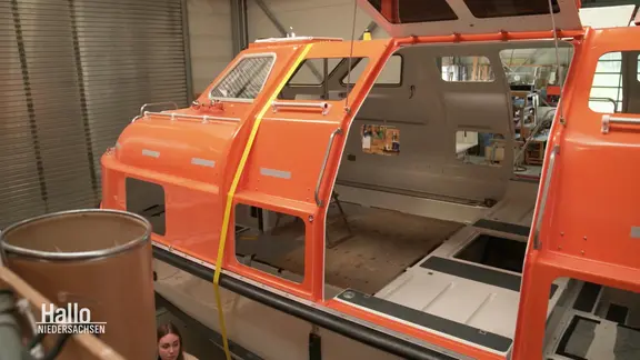 In einer Werkshalle steht ein orangefarbenes Rettungsboot im Rohbau. Eine Frau arbeitet darunter.