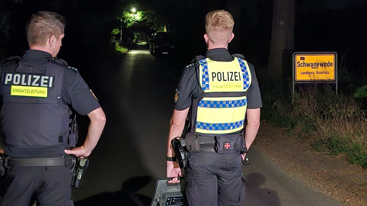 Zwei Polizisten stehen mit einem mobilen Scheinwerfer am Ortseingang von Schwanewede. Im Fall des getöteten Ehepaares in Bremen hat die Polizei den mutmaßlichen Täter festgenommen.