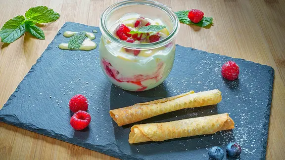 Himbeer-Baiser im Glas mit Waffelröllchen und Beeren auf einer dunklen Schieferplatte