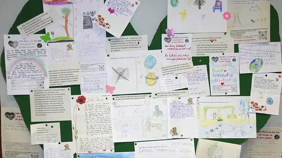 An einer Pinnwand hängen viele Briefe, die meistens bunt gestaltet. Auf ihnen stehen Liebesgeständnisse an den Planeten Erde.