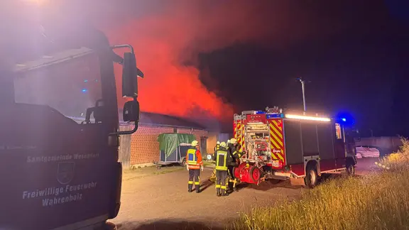 Drei Feuerwehrleute stehen hinter einem Feuerwehrauto. Im Hintergrund steigt aus einem Gebäude Rauch auf.