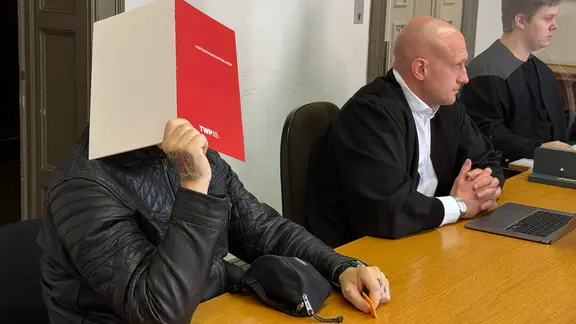 Ein wegen versuchter räuberischer Erpressung angeklagter Mann hält sich im Gericht in Hamburg einen Mappe vor das Gesicht.