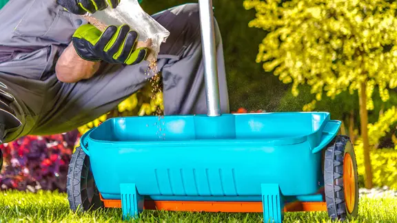 Ein Mann mit Gartenhandschuhen füllt Dünger in einen Streuwagen.