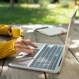 Der Arm einer Frau bedient einen Laptop, der auf einem Tisch in einem Garten steht, während die andere Hand einen Becher hält.