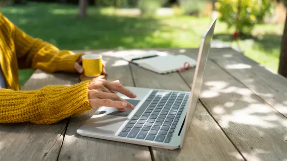 Der Arm einer Frau bedient einen Laptop, der auf einem Tisch in einem Garten steht, während die andere Hand einen Becher hält.