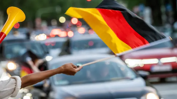 Fußballfans schwenken eine Deutschlandflagge vor einer Straße mit Autos.