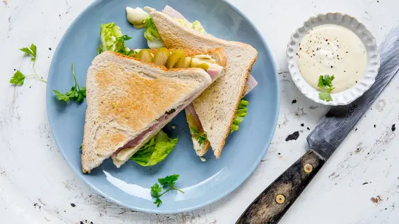 Zwei Käse-Schinken-Sandwiches auf einem Teller mit Honig-Senf-Dip. Daneben liegt ein Holzmesser.
