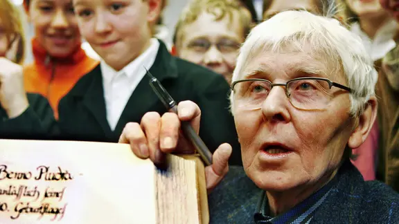 Ο Benno Pludra κρατά ένα χοντρό βιβλίο στην κάμερα. Πίσω του υπάρχουν παιδιά. | εικόνα συμμαχία/ ZB | Νέστωρ Μπάχμαν Ο Benno Pludra κρατά ένα χοντρό βιβλίο στην κάμερα. Πίσω του υπάρχουν παιδιά.