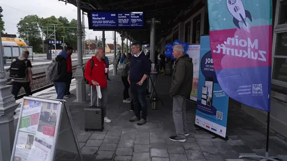 Menschen stehen mit Gepäck an einem Bahnhof.