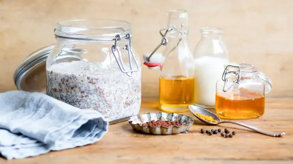 Ein Weckglas mit Overnight Oats, eine Flasche Milch, ein Glas Honig, eine Flasche Leinöl, ein Löffel Kurkuma und ein Schälchen Kakaopulver auf einem Brett.