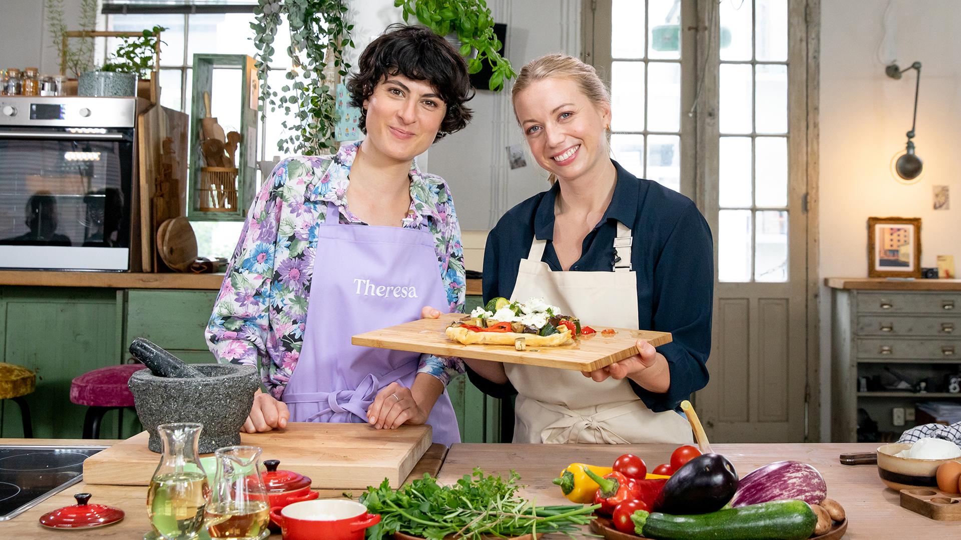 Zora Klipp und Theresa Knipschild stehen mit einem Brett Blätterteig-Pizza in einer Küche. | NDR, Claudia Timmann