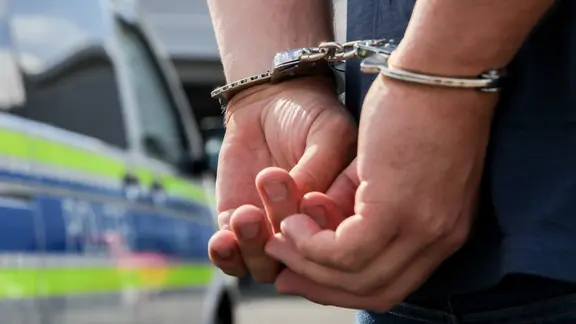 Ein Mann mit Handschellen steht vor einem Polizeiwagen.