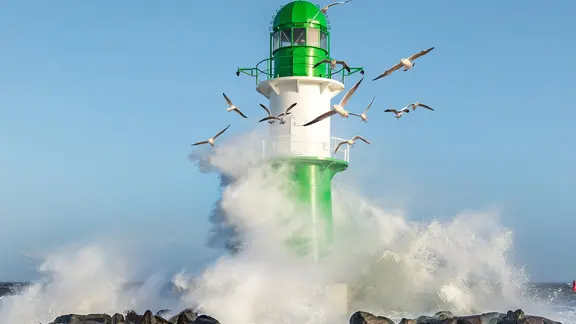 An den Felsen um den Leuchtturm spritzt die Gischt hoch. Um den Leuchtturm fliegen Möwen.