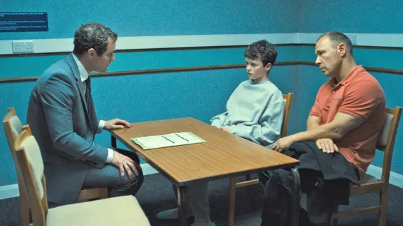 Ein Junge mit dunklen kurzen Haaren sitzt neben einem älteren Mann mit orangefarbendem T-Shirt, beide schauen ernst auf einen Mann im Anzug an der anderen Seite des Holztisches, auf dem Papiere liegen - Szene aus der "Netflix"-Serie "Adolescence"