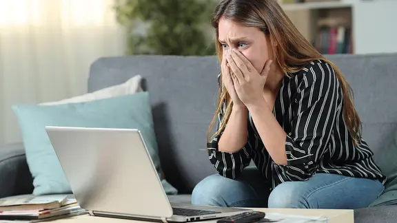 Eine Frau sitzt betroffen vor einem Laptop und hält sich die Hände vor Mund und Nase
