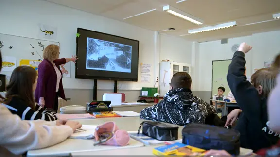 Schüler blicken in einem Klassenraum zur Tafel, eine Lehrerin zeigt aufs Smartboard
