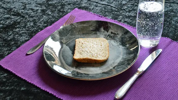 Ein dunkler Teller mit einer Scheibe Brot, daneben Messer und Gabel und ein Glas Wasser | colourbox, Sabine Mederer Ein dunkler Teller mit einer Scheibe Brot, daneben Messer und Gabel und ein Glas Wasser