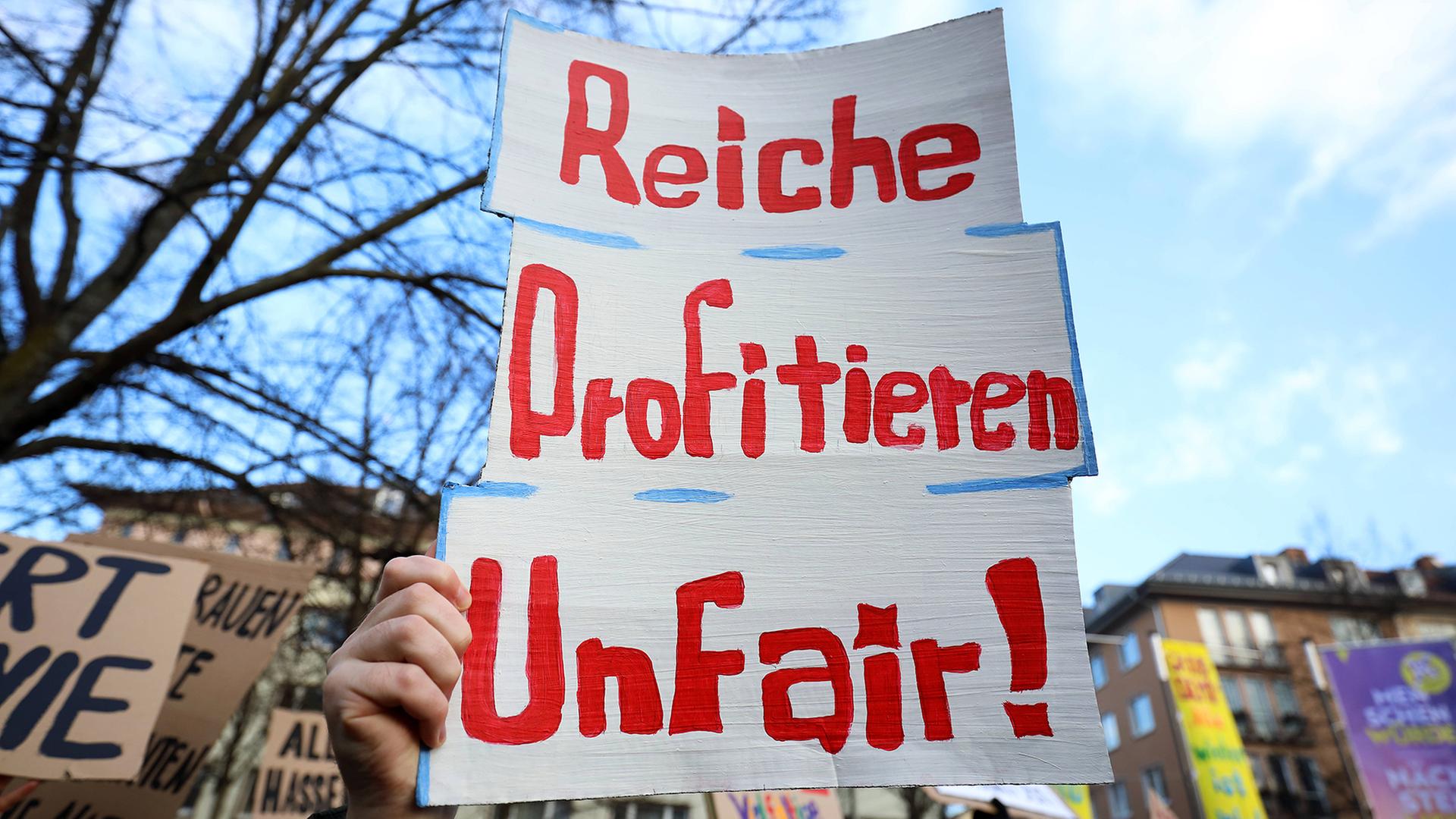 Eine Hand hält ein Schild mit der Aufschrift „Reiche profitieren unfair!“ bei einer Demonstration | Müller-Stauffenberg / IMAGO