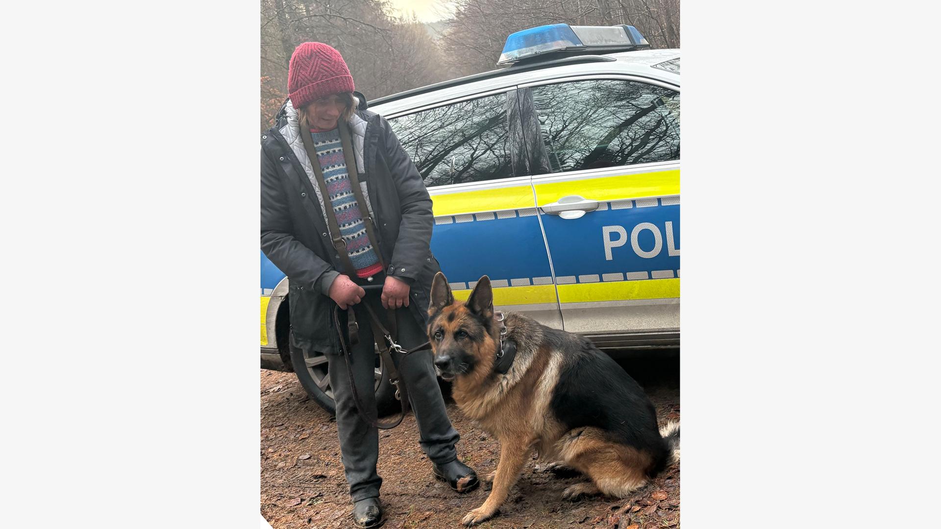 Eine Frau aus Braunschweig steht neben ihrem zuvor vermissten Schäferhund, dahinter Polizeiauto.  | Polizeiinspektion Braunschweig
