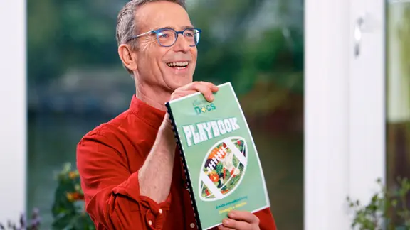 Matthias Riedl hat ein "Playbook" mit Ernährungsstrategien für Fabian W. | NDR/Moritz Schwarz Matthias Riedl hält strahlend ein DIN-A4-Heft namens "Playbook" vor der Brust.