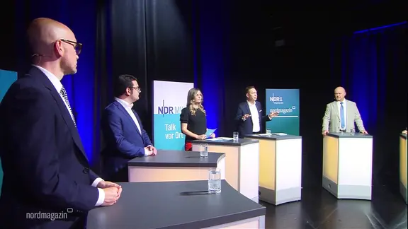 Bei einer Podiumsdiskussion stehen mehrere Vertretende von Partei in einem Halbkreis an Rednerpulten zusammen.
