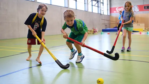 Zwei Jungen und ein Mädchen spielen Hockey in einer Sporthalle.
