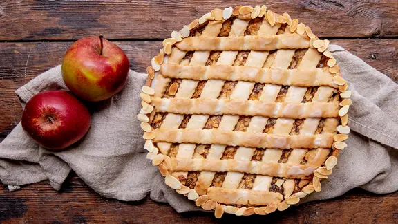 Ein Gedeckter Apfelkuchen steht auf einem Holztisch, daneben liegen zwei Äpfel