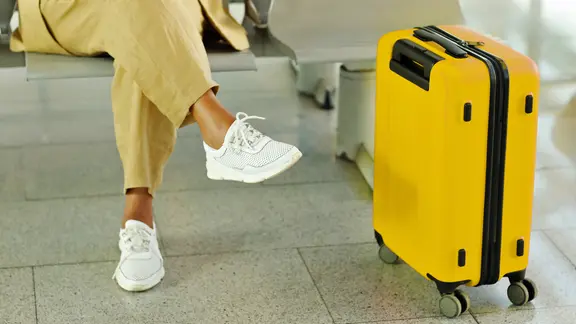 Eine Frau sitzt mit einem gelben Rollkoffer auf einer Bank in einem Flughafen.