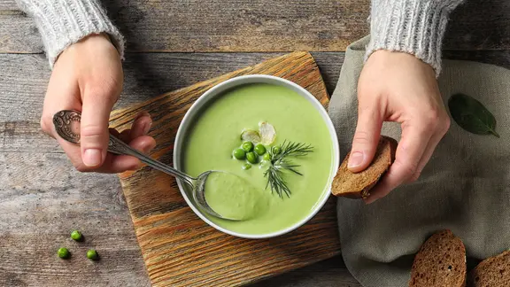 Zwei Frauenhände neben einer Schale mit Suppe, in einer Hand ein Löffel.