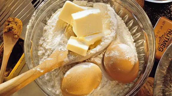 Mehl, Butterstücke, Eier und ein Holzlöffel in einer Schüssel.