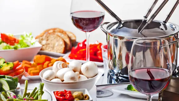 Ein Fondueset, davor Schälchen mit Gemüse und zwei Gläser Wein.