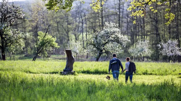 Ein Paar geht Hand in Hand durch eine Parklandschaft mit Bäumen und einer Fuß-Skulptur