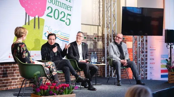 Mehrere Menschen auf einer Bühne im Gespräch, davon links eine Frau, daneben drei Männer.