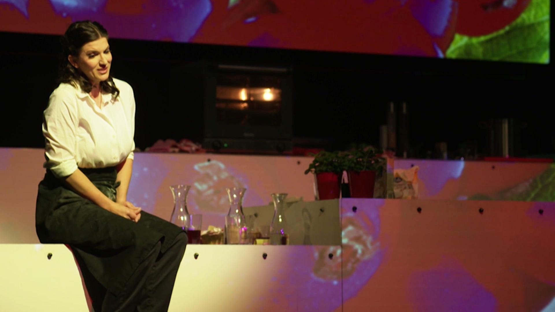 Sängerin sitz auf einer offenen Küche auf der Bühne. | Screenshot