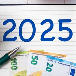 In einem Block steht die Jahreszahl "2025" daneben liegen ein Stift und Geldscheine. Links am Bildrand steht eine Kaffeetasse.