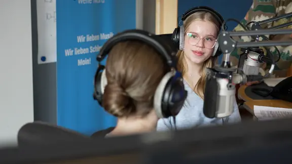 Zwei Mädchen sitzen in einem Hörfunkstudio und zeichnen etwas auf.